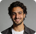 Smiling young man with curly hair and beard wearing a white shirt and dark jacket on gray background.